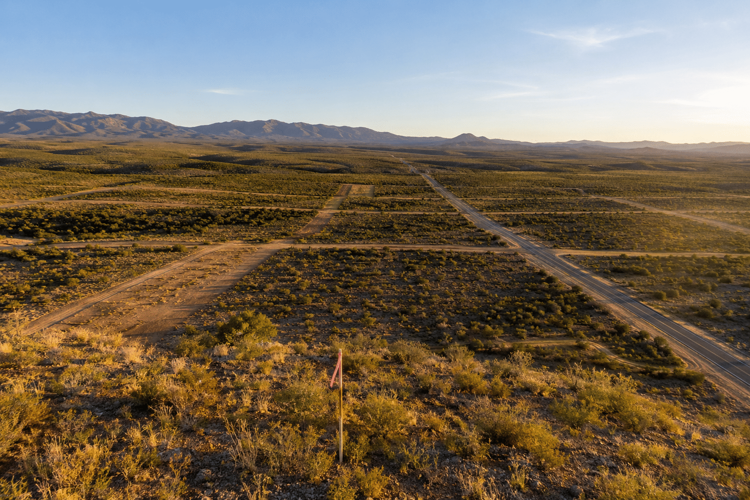 Drone view of undeveloped land in the American Southwest with dirt parcel roads, a paved county road, and a surveyor's stake in the foreground under warm late-afternoon light.