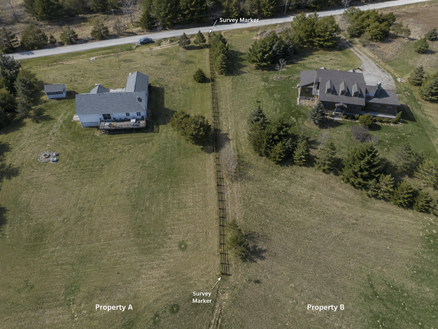 Aerial view of two neighboring rural properties divided by a wooden fence crossing the property boundary, with visible survey markers, open fields, and natural daylight.